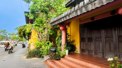 Bright yellow Vietnamese house with red lanterns, lush plants, and motorbikes on the street, ideal...