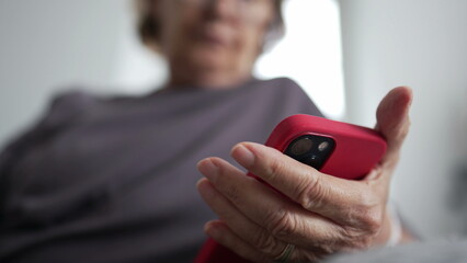 Close-up of a senior woman's hand holding a cellphone while seated on a couch at home, engaged in reading content online