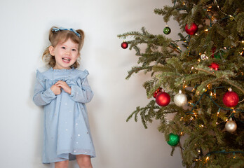 Portrait of a cute Caucasian little girl in a baby blue dress near the Christmas tree. Kids and Christmas concept. Seasonal greetings