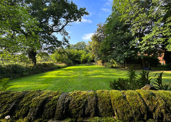 A serene garden with a moss-covered stone wall in the foreground, and lush green grass, trees, and a blue sky. Sunlight filters through the trees, casting long shadows in, Mirfield, UK
