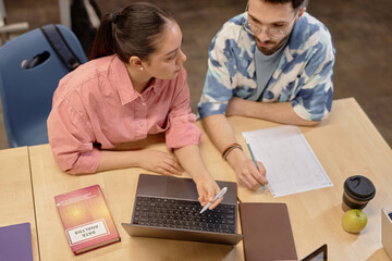 High angle shot of serious Hispanic female student cooperating in studying with male classmate pointing at laptop screen, while sitting at large desk at university classroom