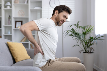 Man sitting on couch holding lower back, experiencing back pain, discomfort or injury. Concept of health issues, chronic pain, posture problems, or muscle strain.