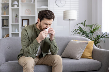 Man sneezing into tissue while sitting on the couch. Indoor scene with man having cold or allergies, showing discomfort and illness.