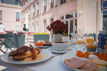 Experience a lavish breakfast spread featuring eggs benedict, cold cuts, fresh fruit, and pastries at a sophisticated hotel with European architecture and lush greenery.