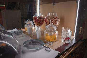 A sophisticated hotel breakfast setup featuring glass jars with dried fruits and granola, fine china plates, silver spoons, and a decorative glass of water with a red flower.