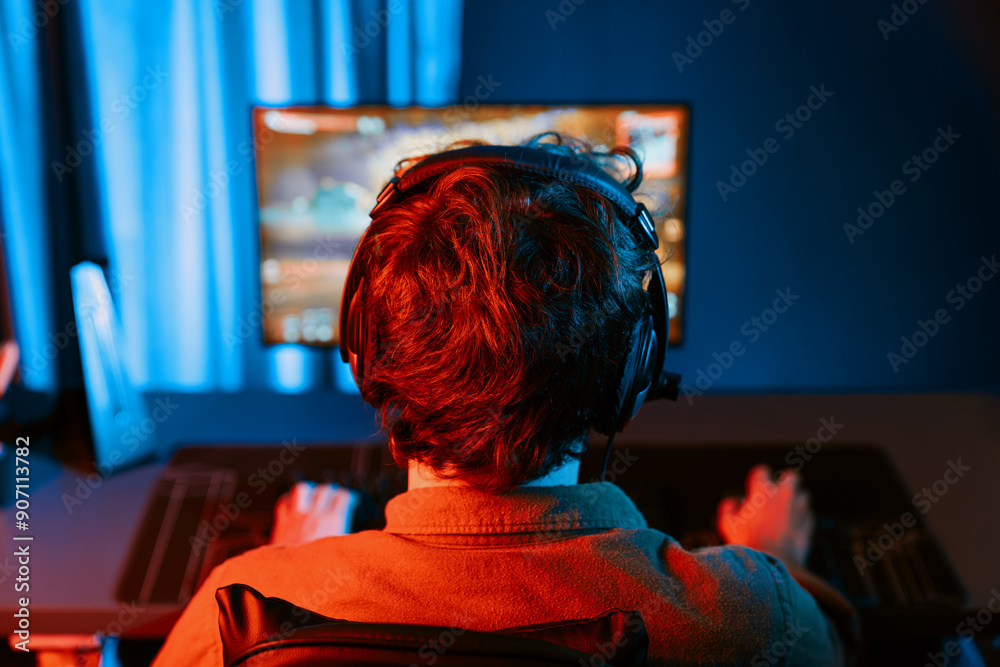 Wall mural Host channel of young gaming streamer, team gamer playing battle game shooting with multiplayer at warship on pc screen with back side image, wearing headset with mic at digital neon room. Gusher.