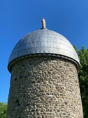 Fieldstone mill with metal roof. Mill tower under a blue sky. Brick tower with old statue of a religious figure from behind on the top.