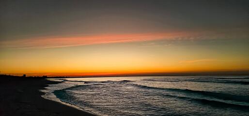 First rays of sunlight break over the sea horizon on a summer day at the beach; landscape view of calm dawn breaking, sunrise over Mediterranean coast with pink clouds and shallow ocean with waves.