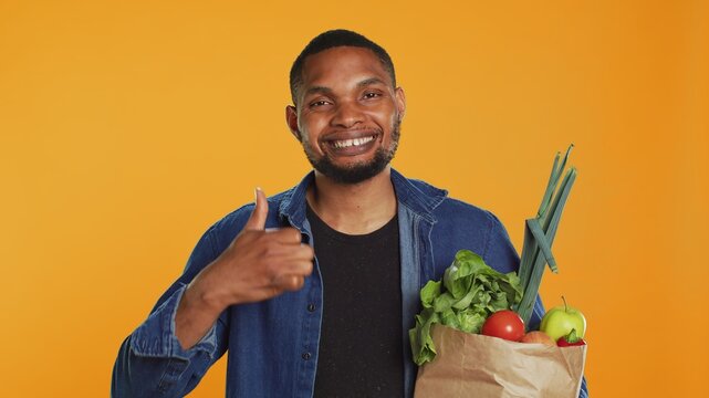 Cheerful positive guy giving a thumbs up for locally grown produce, advocating for zero waste and healthy eating from farmers market. Young adult doing a like gesture in studio. Camera A.