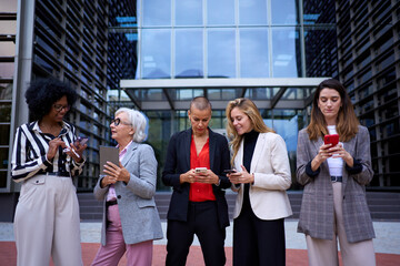 Team of multicultural business women in formal wear standing using and looking mobile phone. Focused adult professional females with cells in hand outdoor. Technology addicted concept 