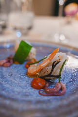 A beautifully plated gourmet seafood dish featuring shrimp and octopus, garnished with green vegetables and orange sauce, set in an elegant luxury hotel restaurant.