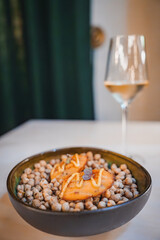 Sophisticated chickpea dish topped with garnished appetizers, served with white wine. Dark green curtain backdrop adds elegance, indicating a high-end restaurant or luxury hotel.