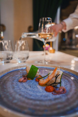 Beautifully plated seafood dish with vibrant vegetables and herbs on a blue ceramic plate. A hand pours white wine, enhancing the elegant dining experience.