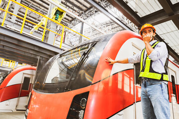 Asian male handsome operator operator engineer standing in front of electric locomotive at modern electric train maintenance station checking safety systems before using city transportation service.