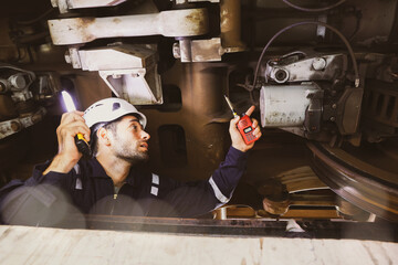 Male train maintenance engineer uses light to look at the undercarriage and check the safety of the electric train brake control system in a station repair garage and report to the repair team.
