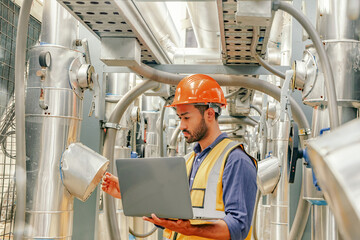 Male HAVC refrigeration system technician stands holding laptop recording preventative inspection...