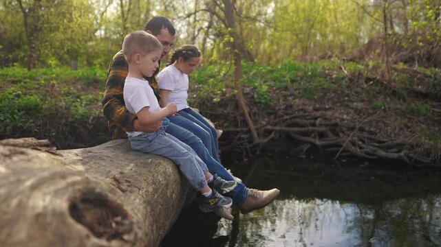 dad with children sits on a log. happy family child dream concept. happy young dad sitting with his son and daughter in the forest on a large log above the river lifestyle. kids dad's