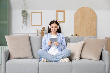 Beautiful young Asian woman in headphones listening to audiobook with modern tablet computer and sitting on sofa in living room