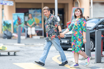 50-something Malay man and 30-something Chinese woman couple in colorful clothes walking across a crosswalk in Chinatown Kuala Lumpur, Malaysia.