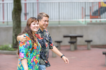 Malay man in his 50s wearing colorful shirt and Chinese woman in her 30s, walking closely together in KLCC Park in Kuala Lumpur, Malaysia.