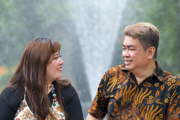 Malay man in his 50s wearing traditional Malaysian shirt and Chinese woman in her 30s wearing traditional dress, sitting closely together in front of fountain in Taman Botani Perdana in Kuala Lumpur