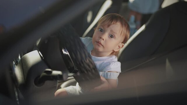 a small boy sits behind the wheel of a car. people kids and games concept. a little kid plays behind the wheel of his parents' car, holds and lifestyle turns the steering wheel