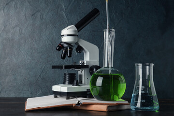 Modern microscope with chemical flasks and books on wooden table against grey grunge background