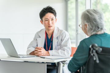 Obraz premium 病院・介護施設で高齢者女性を真剣な表情で診察・問診する若いアジア人男性医師 