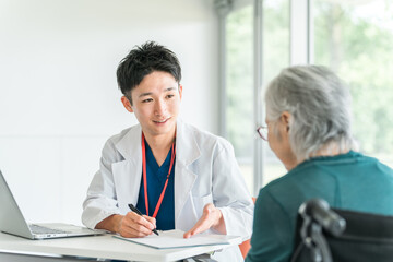 病院・介護施設で高齢者女性を笑顔で診察・問診する若いアジア人男性医師（かかりつけ医）
