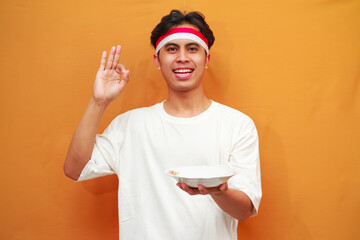 Happy Indonesian man holding empty plate on yellow background.