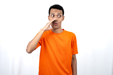 Portrait of a young Asian man wearing an orange t-shirt with his hand near his mouth whispering gossip and telling secrets while sharing news isolated on white background.