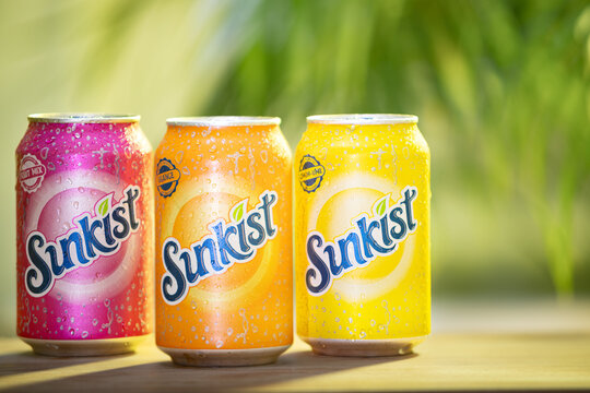 Vibrant and refreshing cans of Sunkist soda in different flavors: fruit mix, orange, and lemon-lime which are covered in condensation droplets on outdoor setting perfect for enjoying a cold beverage.