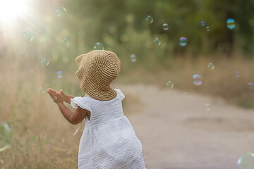 baby and soap bubbles. A little girl seen from behind in a white dress and a straw panama hat walks on a summer day and happily catches soap bubbles