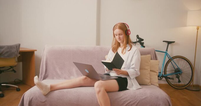Woman in earphones types on laptop holding book in hand