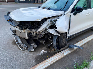 A white car with a damaged front end is parked on the side of the road, possibly due to a collision. The wheel, tire, hood, and grille seem affected, indicating significant damage