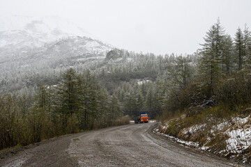 A truck drives on a dirt road on a mountain pass. Cold snowy weather in early June in the Far North. Summer snowfall in the mountains. Beautiful landscape with a winding road. Magadan region, Russia.