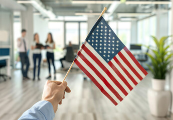 Close up of an American flag being held in the style of one person in the foreground, blurred people holding US flags and dressed in business casual standing behind him in a blurry background.
