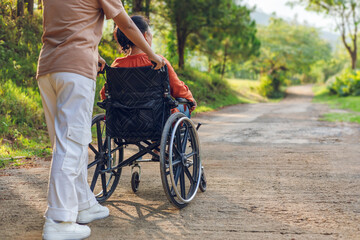 Obraz premium Woman sitting in wheelchair with his care helper walking on park trail outdoor morning sunrise mountain background. Rear view