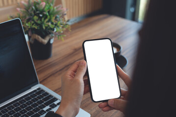 mockup blank screen cell phone for web template or mobile app design. Woman hand using mobile phone during working on laptop computer on wooden table at coffee shop or home office