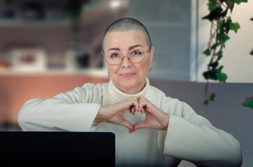 Elderly woman with glasses, in a white turtleneck, smiles and forms a heart with her hands, capturing a moment of joy and connection while working from home.