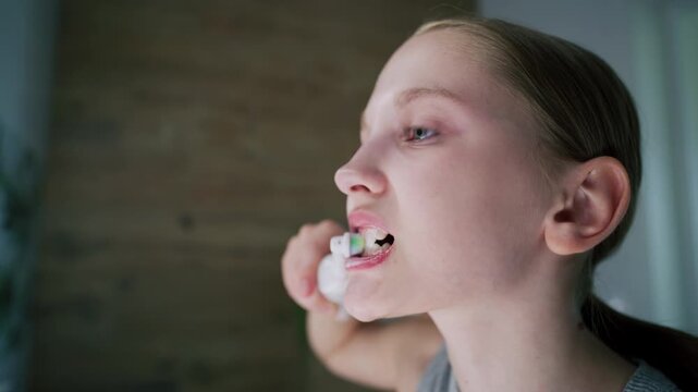 Close up of young girl looking at mirror in bathroom, brushing her teeth. Pretty teenager stands in front of sink, uses electrical toothbrush before bedtime. Evening bath routine. Lifestyle concept.