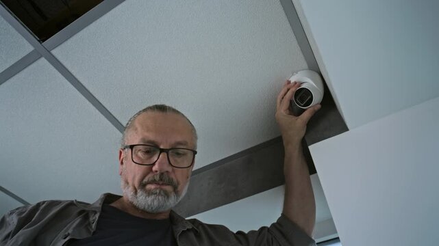 Male mature technician sets up the angle of CCTV camera in office, adjusts it using tablet computer with professional application. Monitoring and tracking system. Surveillance, security and privacy.