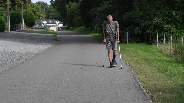 man with broken leg walks down the street,his left leg is in cast adapted for walking in special shoes,he moves with crutches,which reduces the load on the broken foot,concept of rehabilitation