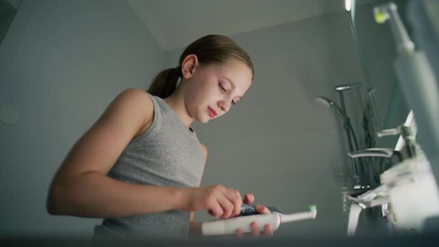 Attractive young girl looks at mirror in bathroom, brushes her teeth. Pretty teenager stands in front of sink and uses electrical toothbrush before bedtime. Evening bath routine. Concept of lifestyle.