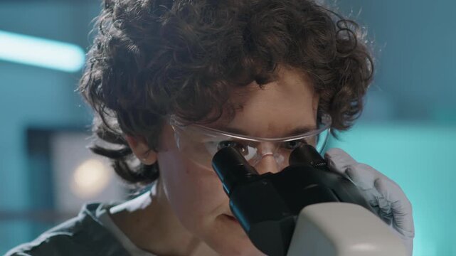 Close-up shot of face of curly female laboratory technician or pathologist in transparent protective goggles, blue medical uniform, gloves looking at tissue sample into microscope and adjusting focus