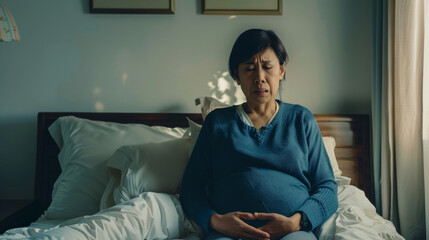 Obraz premium Pensive pregnant woman sitting on a bed, cradling her belly, in a softly lit room filled with serenity and anticipation.