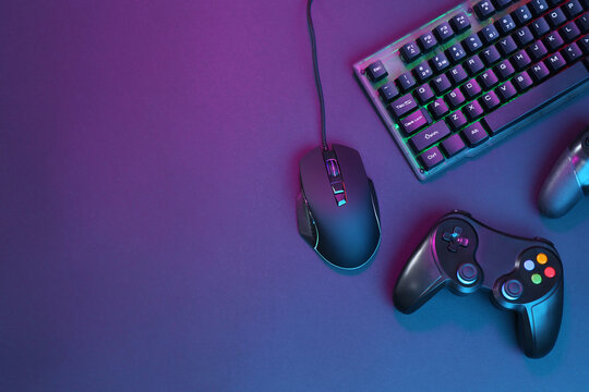 Computer mouse, RGB keyboard and game controllers in neon lights on dark background, flat lay. Space for text