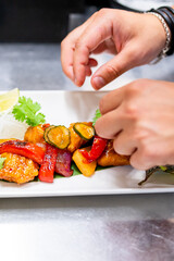 Close-up of a hand garnishing a colorful plate of grilled vegetables with a slice of lemon, highlighting fresh and healthy cuisine