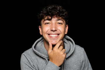 Portrait of young man about 25 years old with curly hair, holds hand on chin and smiles at camera. Isolated on black studio background