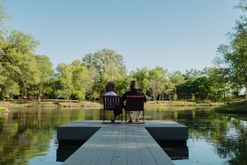 Obraz premium Back shot of couple sitting on chairs at the end of a dock overlooking a serene lake on a sunny day